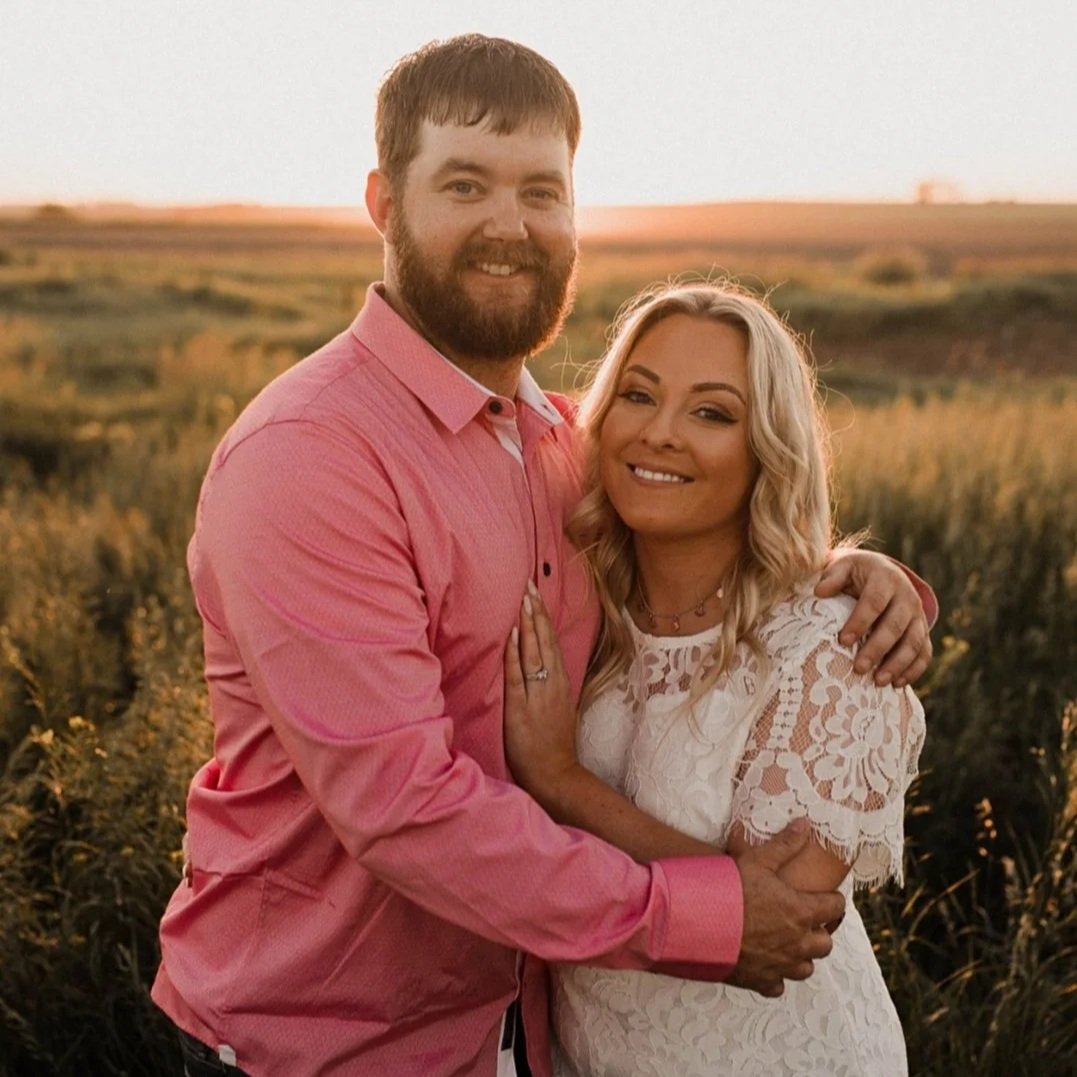 Couple embracing in a sunlit field during sunset, showcasing romantic outdoor engagement photography. The man wears a pink button-up shirt, while the woman is dressed in a white lace dress, highlighting a warm and intimate moment.