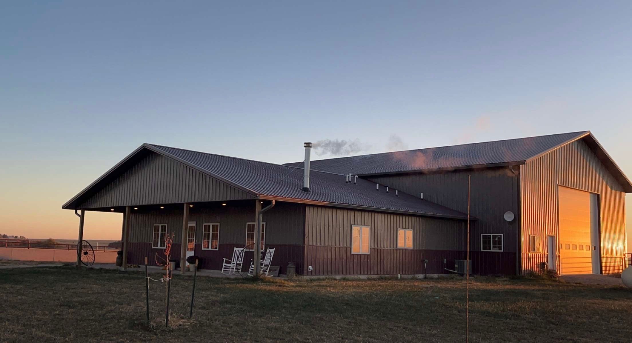 Modern farm building at sunset with a metal roof, featuring a porch, windows reflecting warm light, and a smoke chimney, surrounded by open fields.