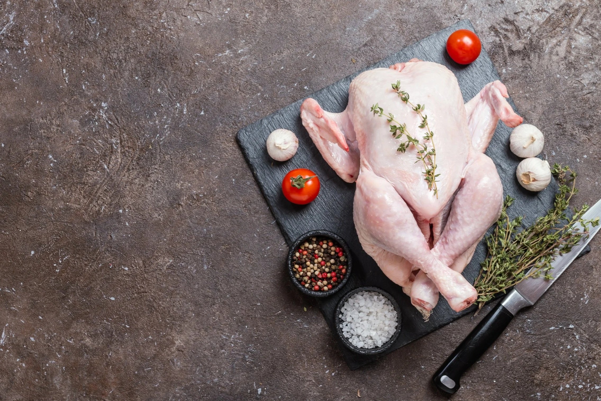chicken_whole_raw Raw whole chicken on a black slate cutting board, garnished with fresh thyme, surrounded by garlic, tomatoes, and bowls of mixed peppercorns and sea salt, with a kitchen knife nearby.