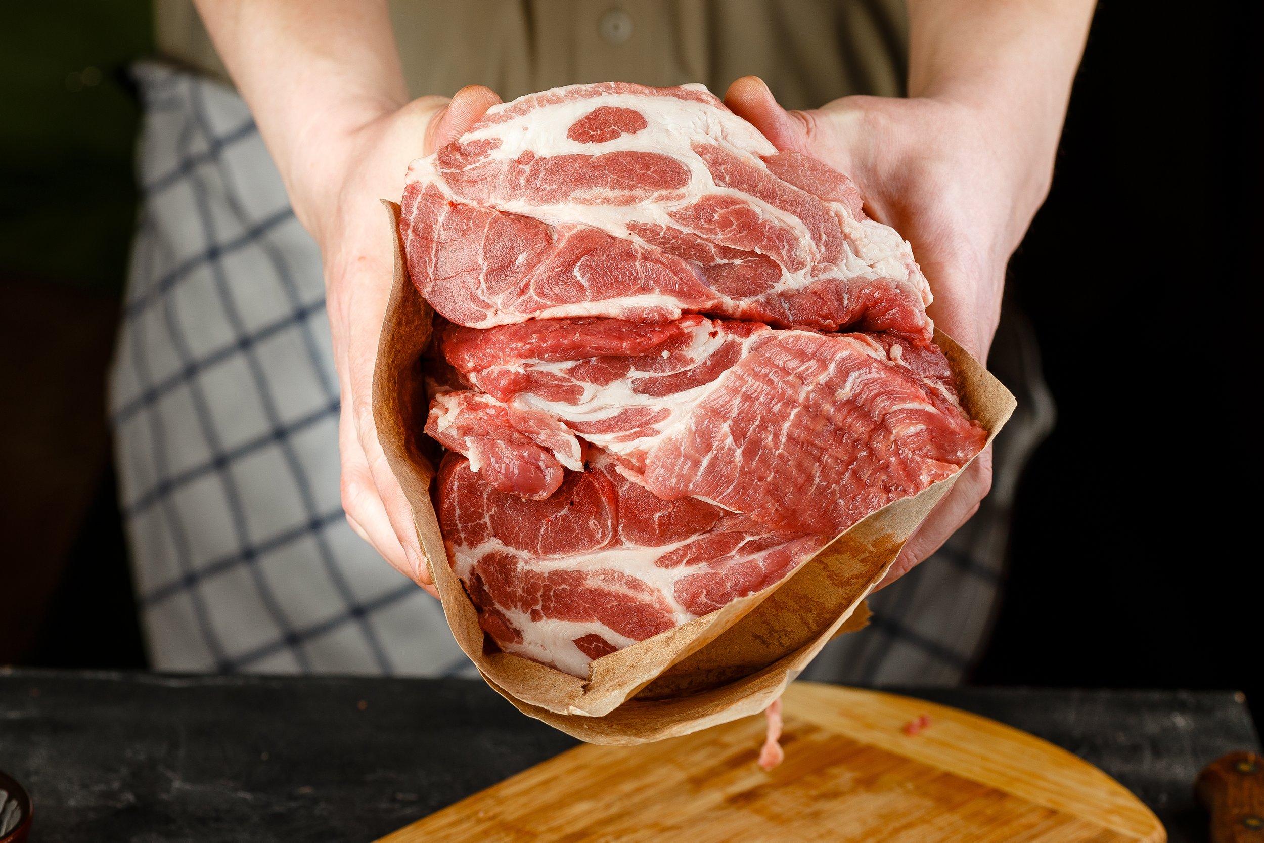 Close up of Male hands chopping knife pork Fresh cuts of pork meat held in brown butcher paper, showcasing marbled texture and quality, with a wooden cutting board in the background. Ideal for cooking enthusiasts and chefs.