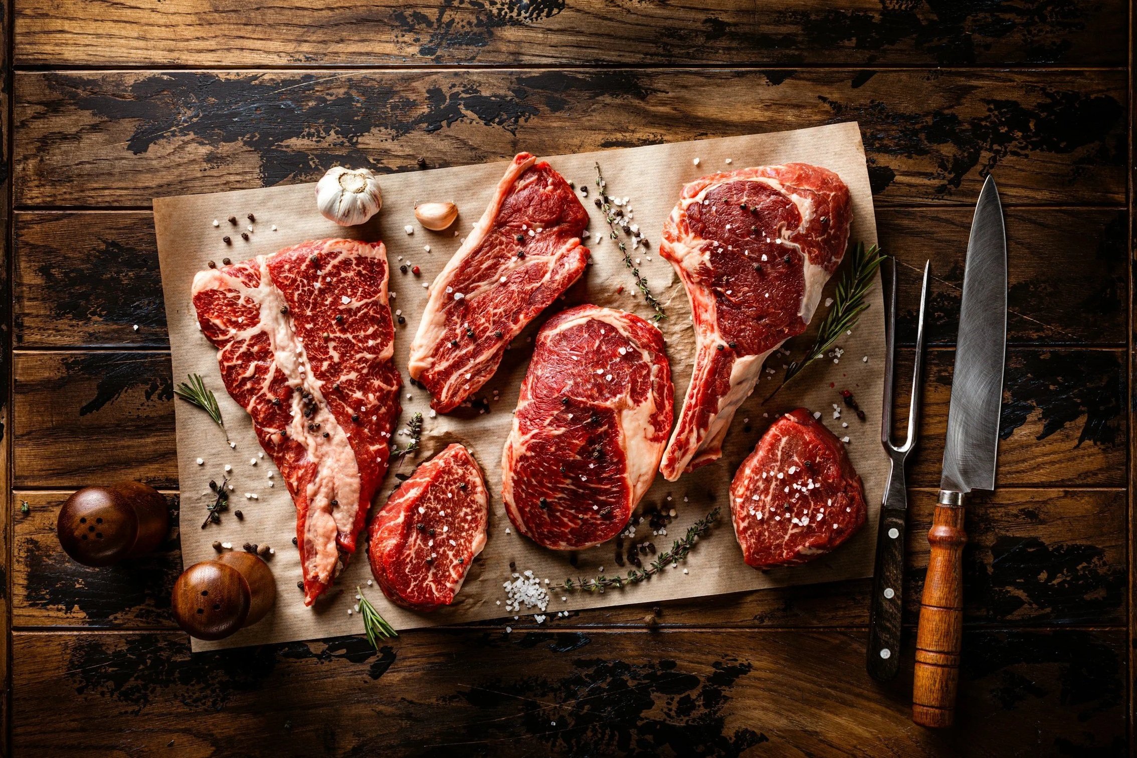 unsplash-image-JHWfH8V2INk Assorted cuts of marbled beef displayed on parchment paper, garnished with garlic, herbs, and spices, alongside a carving knife and fork on a rustic wooden surface.
